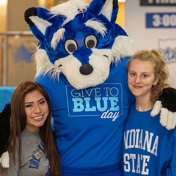 Sycamore Sam stands with arms around two smiling female students, a taller blonde woman wearing a blue Indiana State sweatshirt on the right and a shorter Latina woman wearing a gray Indiana State Sycamores on the left. To their right are blue and white balloons. Sycamore Sam is wearing a Give to Blue t-shirt.