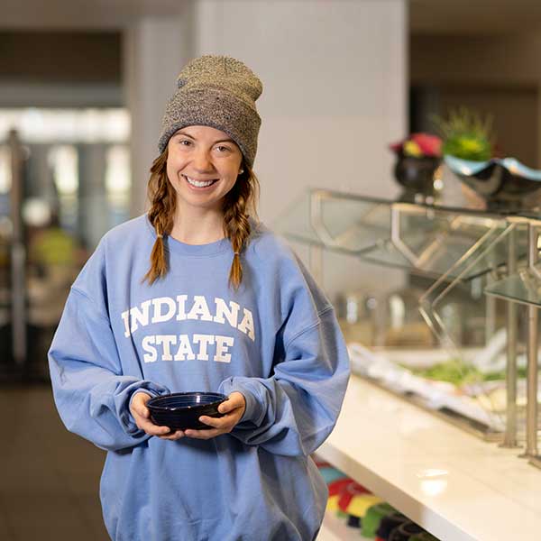 A smiling female student in a grey wool hat and a blue Indiana State sweatshirt holds a bowl next to the salad bar in the dining hall.