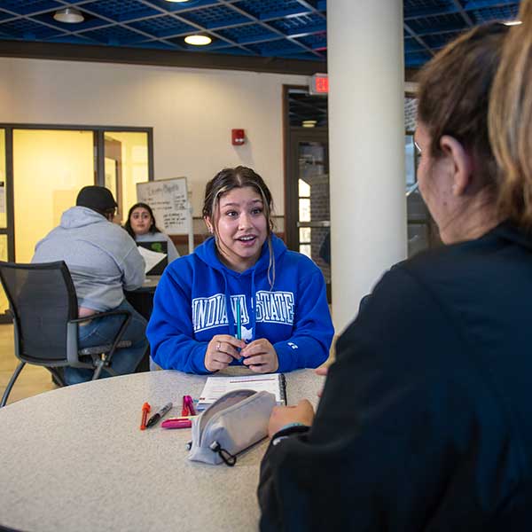 A student in a royal blue Indiana State sweatshirt is seated at a table in a commons area, speaking to another student who is mostly unseen. Two other students are visible at a table behind them.