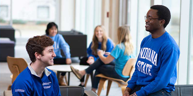 Two students wearing blue Indiana State shirts laugh together in the Commons areas of a residence hall. Several other students are seen sitting and smiling in the background.