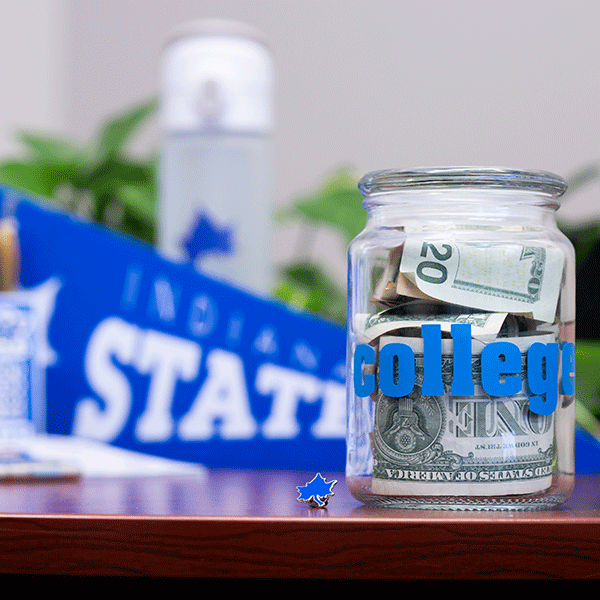 A photo of a jar that has cash in it and says “college” on the front of it. Indiana state gear is blurred in the background. 