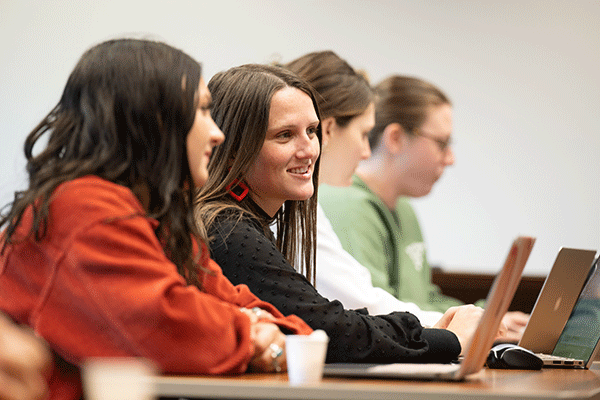 A white female student with dark brown hair sits in a class. Visible to her left, slightly out focus, sits another smiling female student with brown hair and a laptop. In the background out of focus, two other women are visible, one with dark hair and a white shirt, the other with light brown hair and glasses.