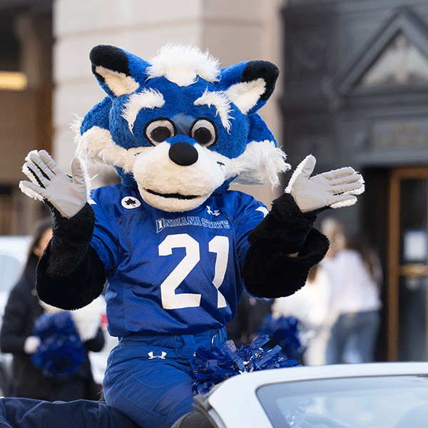 Sycamore Sam, Indiana State’s official mascot, sits on the backseat of an open-roof car during a parade. He wears a blue Indiana State football jersey, and he is blue-and-white colored. A building is blurred in the background.
