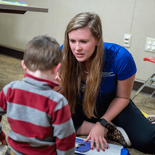 A white female student with long blond hair is sitting on the ground with a toddler boy with short brown hair. The female student wears a blue Indiana State University T-shirt, blue jeans, and cheetah-print shoes. The toddler wears a red-and-grey long-sleeved shirt.