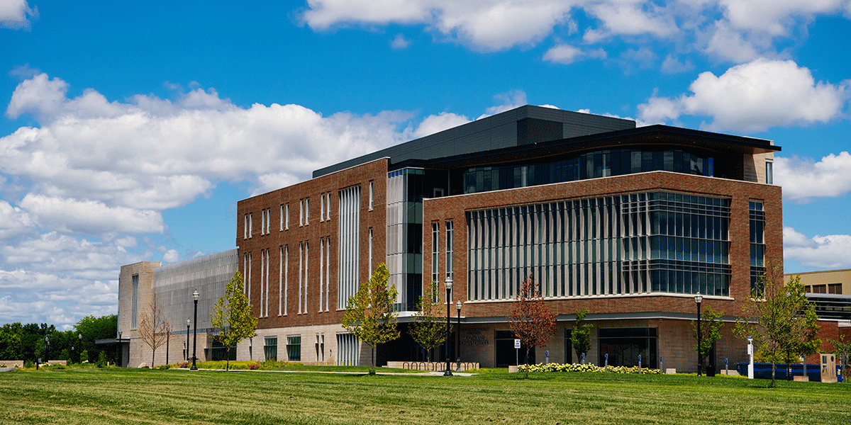 An exterior view of the College of Health and Human Services building, a brick building with numerous glass windows on the sides of the building. A green lawn is in front of the building.
