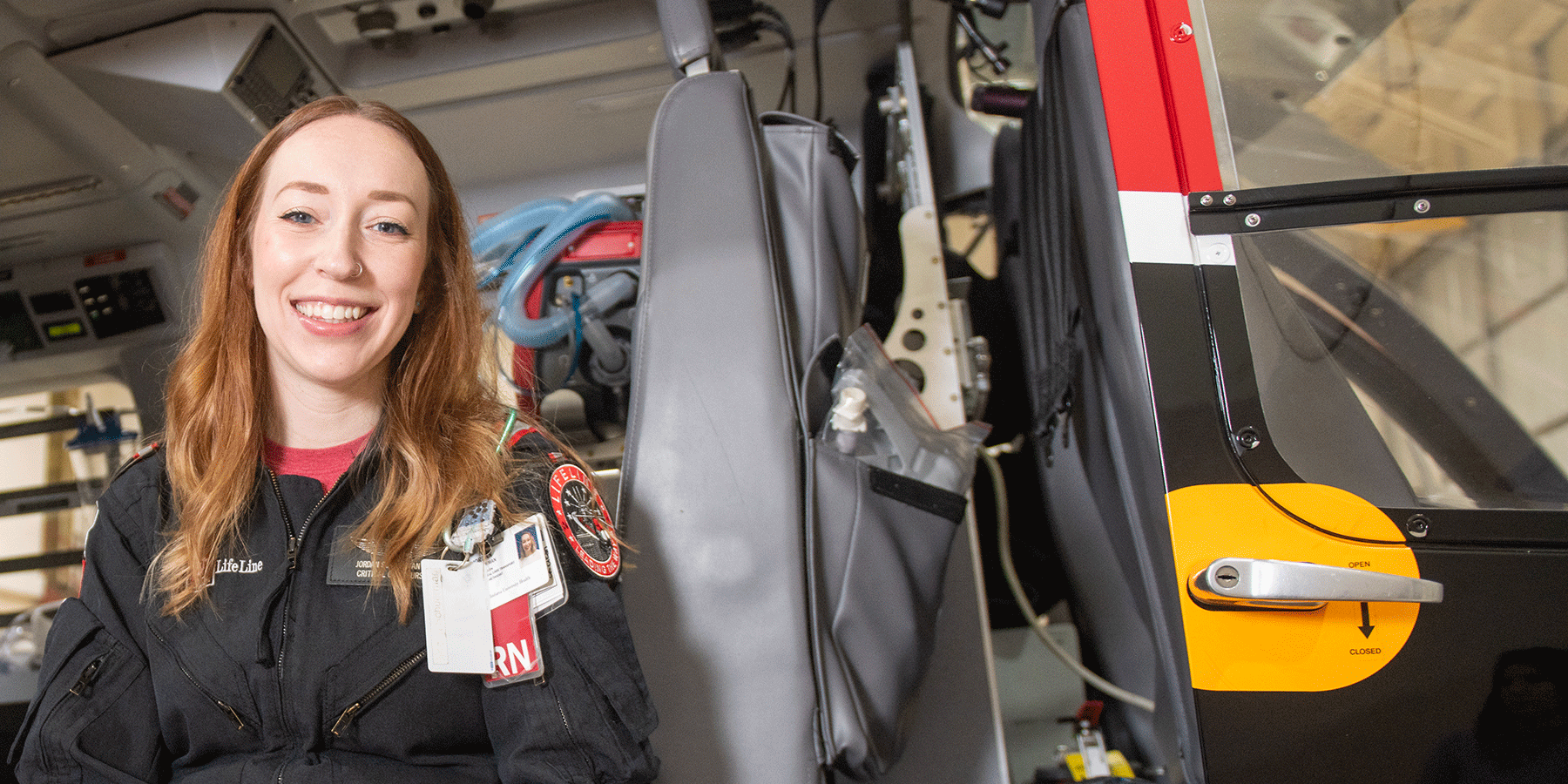 A white female student poses in an emergency services LifeLine vehicle. She has long red hair, a nose ring, and she wears a back LifeLine uniform with a red undershirt. A badge is on her chest. Medical devices and equipment are in the vehicle.
