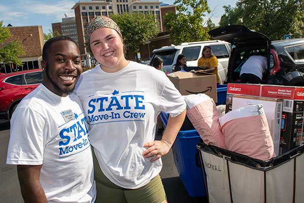 To students in white Move-in Crew T-shirts pose next to a cart of residential room belongings