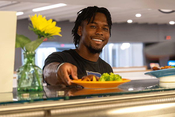 A Black male student smiles as he is handed a plate in line at the Sycamore Dining Hall.  