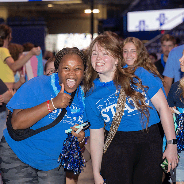 Two female students in blue ISU shirts smile, one is giving a thumbs up