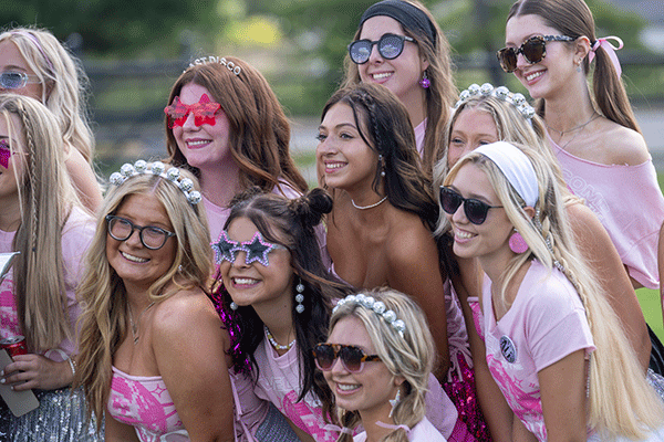A group of female students in a sorority with pink shirts and a variety of styles of sunglasses pose for a photo at an outdoor event.