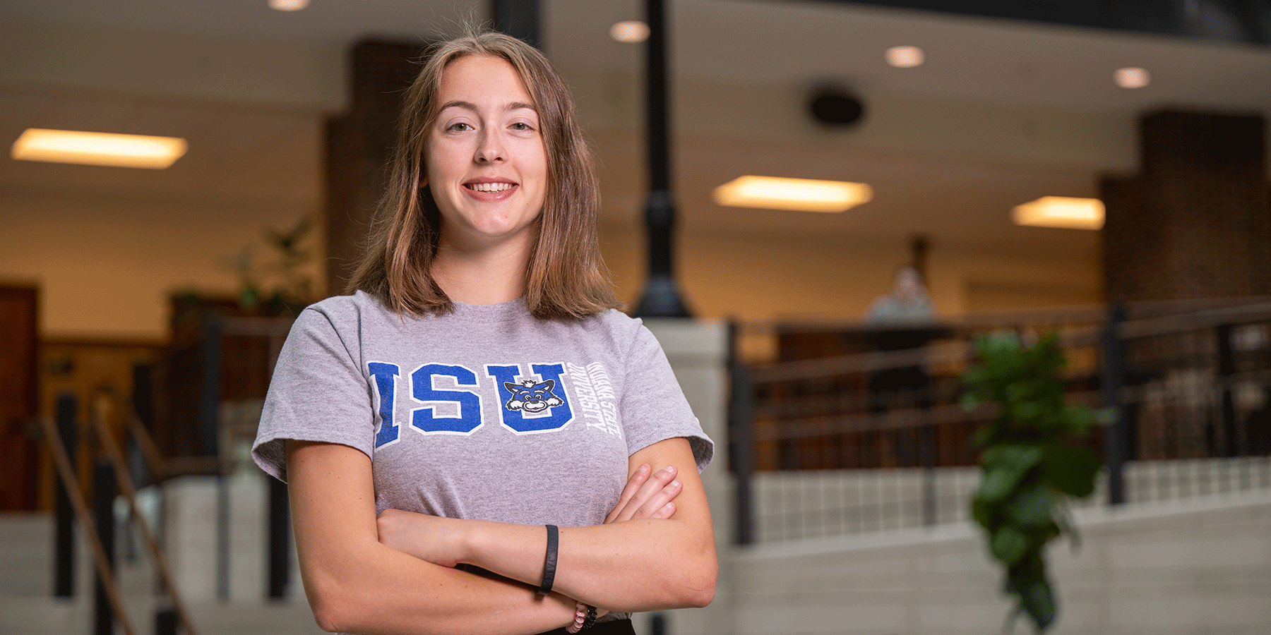 A female student with shoulder-length brown hair. She wears a grey-and-blue ISU T-shirt, and she poses with her arms crossed in an atrium.
