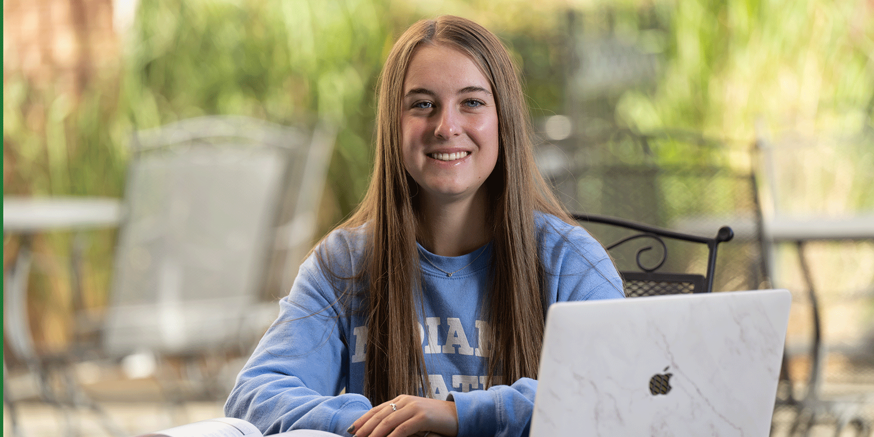 A female student with long brown hair. She wears a blue Indiana State sweatshirt. She sits at a table outside with a white laptop and an open textbook.