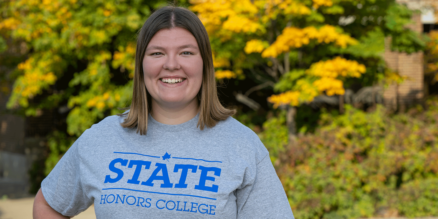 Honors student Kaylan Galey, smiling, with shoulder-length brown hair, is wearing a gray Indiana State Honors t-shirt. Trees with green and yellow leaves are visible in the background.