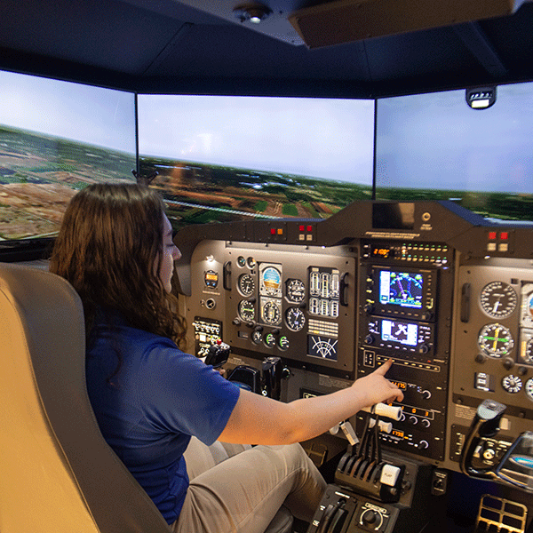 A student with dark hair, wearing a blue shirt and beige pants, works the controls of a flight simulator. An aerial scene is visible on the three simulation screens. The simulator control panel features switches, buttons, gauges, and digital screens.