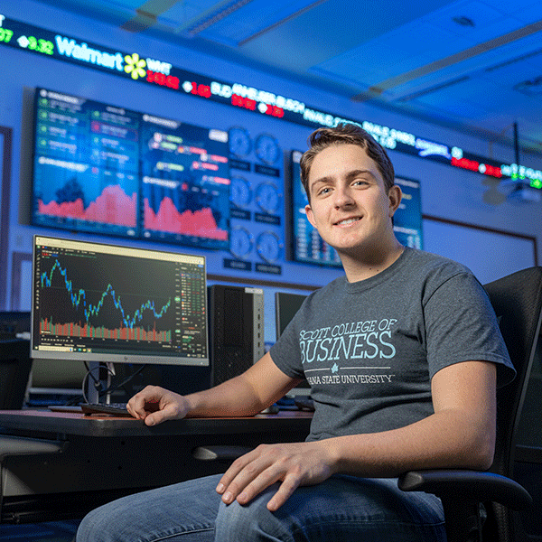 An Indiana State business student with short brown hair and wearing a gray Scott College of Business t-shirt sits next to a computer in the Scott College of Business virtual trading floor. The computer is displaying graphical stock data. On the wall behind, large monitors display more detailed stock data. Above that, an electronic stock ticker shows current trading values – Walmart is the one visible company name. Soft, electric blue light illuminates the scene.