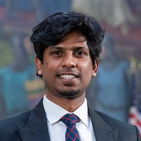 A man with tanned skin and black hair poses. He wears a black dress suit, a white dress shirt, and a blue-and-red tie. Blurred in the background is an American flag and a mural.