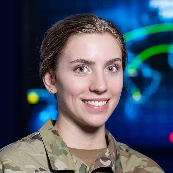 A woman with dark blonde hair poses. She wears a camouflage military uniform. Blurred in the background is a digital map with green lines.