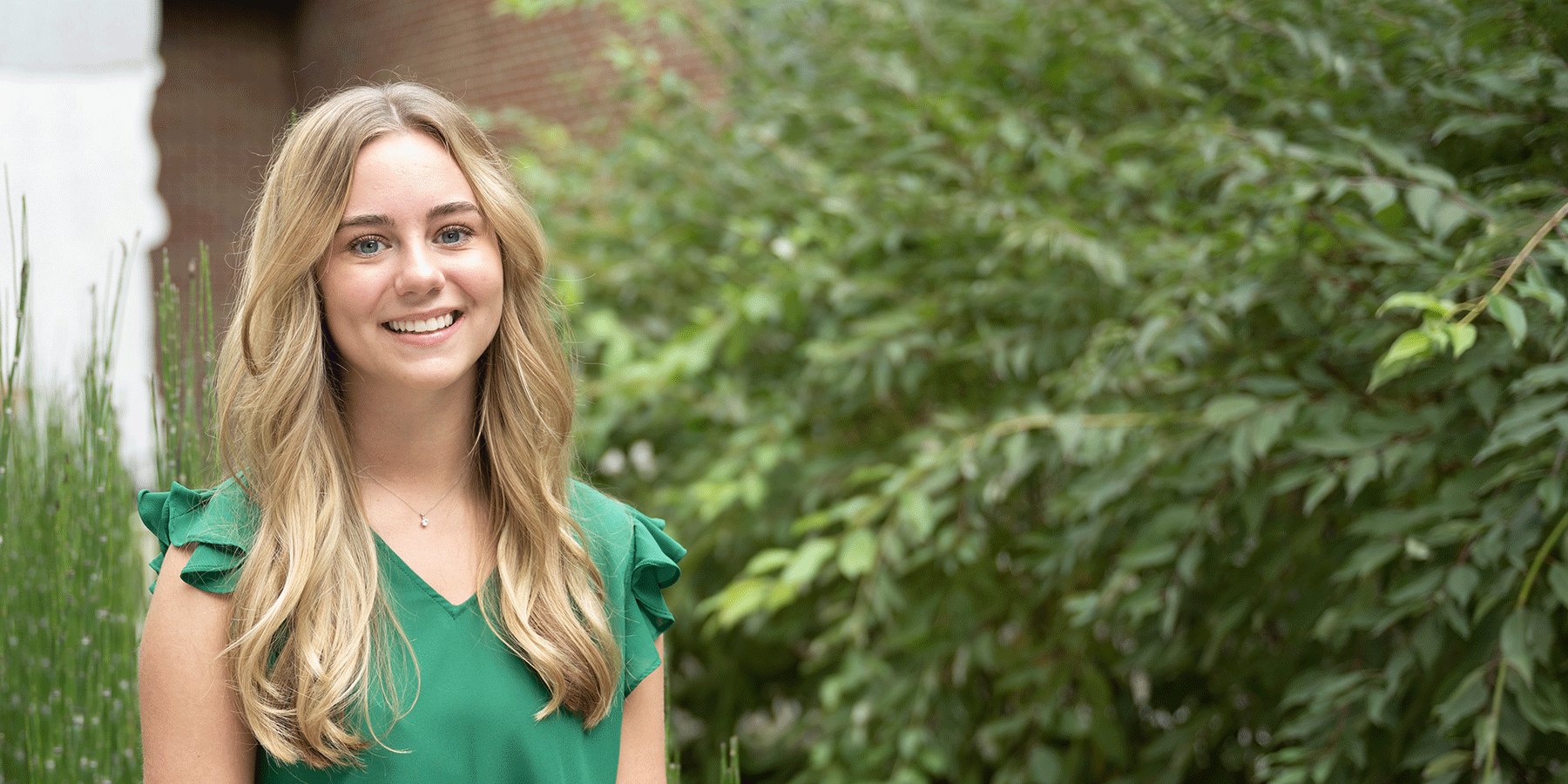 A smiling, blonde, female student wearing a green, shoulder-sleeve blouse is shown outdoors on campus, with green leaves and grasses in the background and to the right of the image.