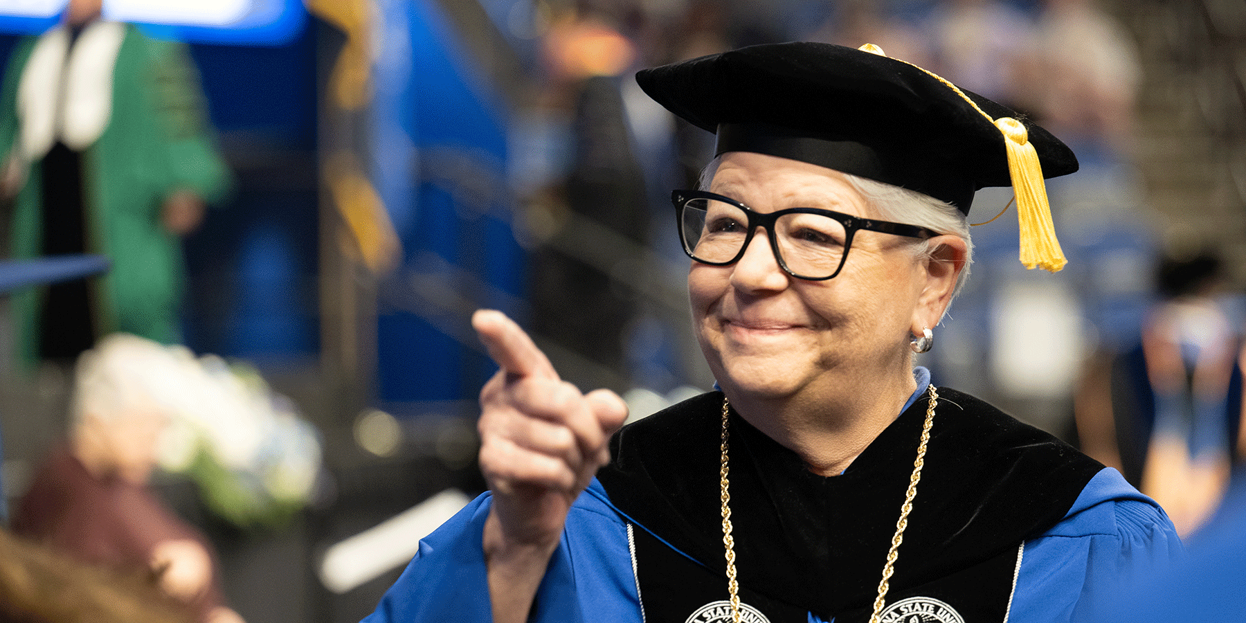 A smiling President Deborah J. Curtis speaks with two students who are visible from the back. Dr. Curtis wears glasses and a black-and-white sweater over a royal blue turtleneck. Ceiling lights are blurry but visible in the background.