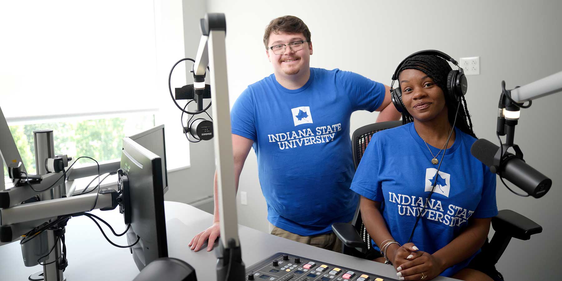 A White male student and a Black female student smile as they stand in the Indiana State radio station. Both wear blue Indiana State t-shirts. Microphones, a computer, and control board are also visible.