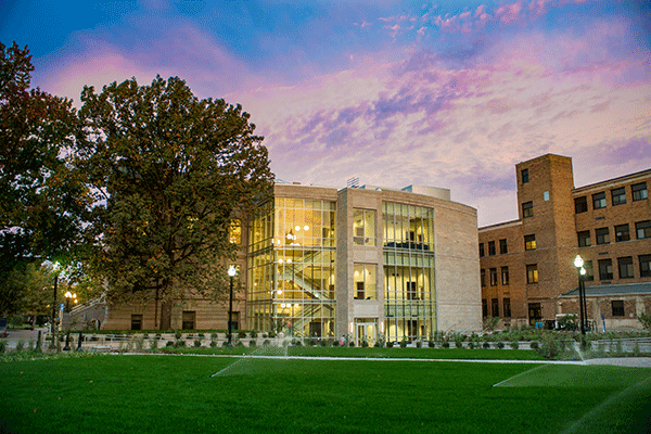 The Fine Arts Building at Indiana State is illuminated at a distance through its many glass windows beneath a pink and purple evening sky.