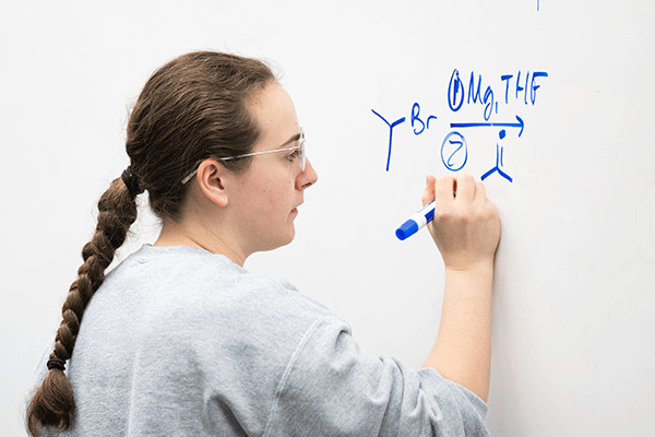 A student writes a chemical formula on a whiteboard with her blue marker.