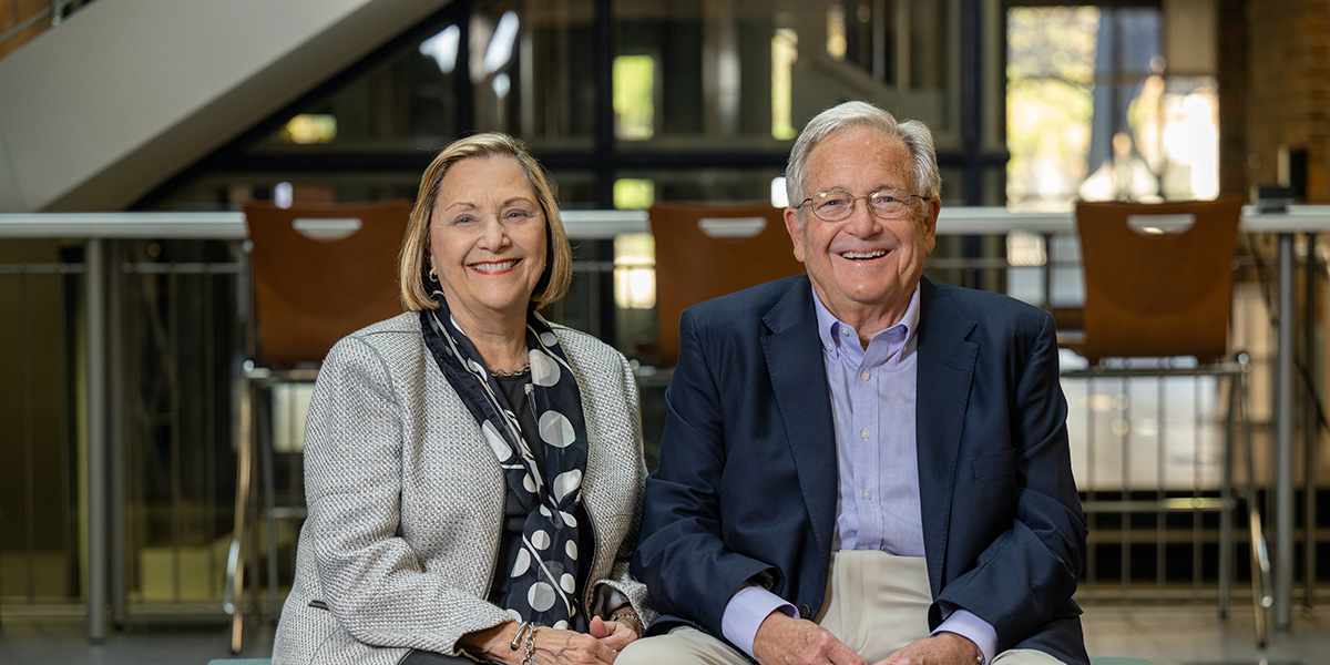 A smiling couple, Gloria and Steve Bailey, seated in the Scott College of Business. Steve is wearing a light blue shirt, a dark blue jacket, and beige pants. To his right, Gloria is wearing a business casual white jacket with dark blouse and pants and a black scarf with large white circles on it.