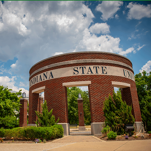 The circular entryway arch at Indiana State, made of red and white brick and with the words Indiana State visible along the top. Green trees and shrubs are also visible.