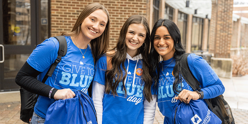 Three smiling female students stand outside the Hulman Memorial Student Union. All three wear Give to Blue Day t-shirts. All three have long, dark hair. The one on the right and on the left are holding blue drawstring book bags.