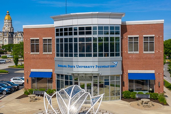 Aerial shot of the two-story Indiana State University Foundation building. Red brick is visible on the sides with a white and silver center façade featuring glass windows and doors. The Foundation’s name in blue letters appears between the first and second floors. There is a large Plexiglass sculpture of a Sycamore leaf in front of the building. The Vigo County Courthouse is visible in the distance on the left.