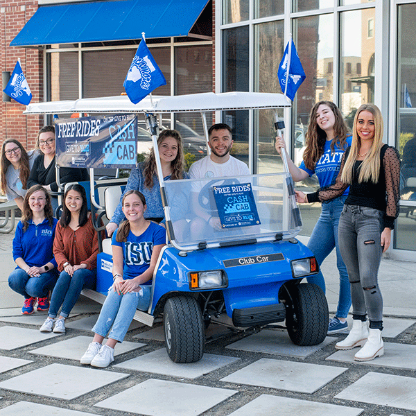 Nine Indiana State students, sit on or stand around the Cash Cab, a golf cart decorated with blue signage advertising free rides as part of Give to Blue Day. There is one male student in the image; he is seated behind the wheel of the golf cart. Several students are wearing blue Indiana State shirts.