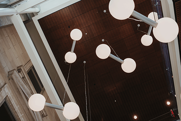 Art installation featuring white and gray art pieces that look like barbells, suspended from the ceiling in the Health and Human Services Building. The five sets of round white end pieces and the gray connectors stand out against the dark ceiling above.