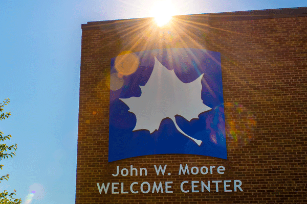 A large white Sycamore leaf logo on a blue background is visible above the name of the John W. Moore Welcome Center. The sun is peeking just above the red brick building, casting rays of sunshine over the top of the logo.