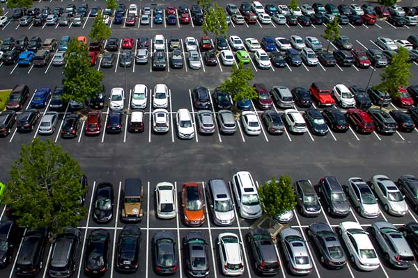 Aerial shot of a large parking lot filled with cars. Occasional green trees can be seen around the lot.