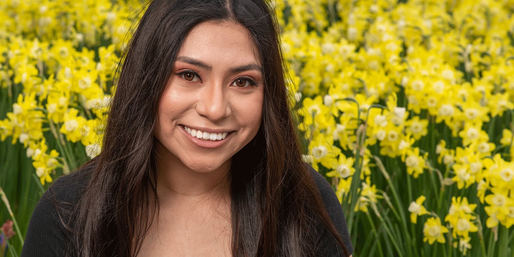 A smiling young Latina woman with long, dark hair is shown with a background of yellow and white flowers.