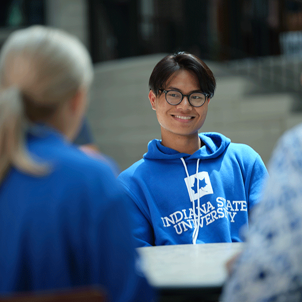 A young Asian American man with black hair and glasses smiles and chats with a blonde woman sitting opposite him. He is wearing a blue and white Indiana State University hoodie.