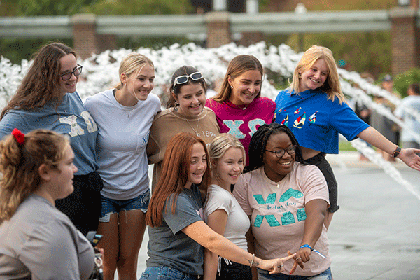 Nine students of mixed races pose in front of a fountain. They wear a variety of grey, blue, brown, and pink clothing.