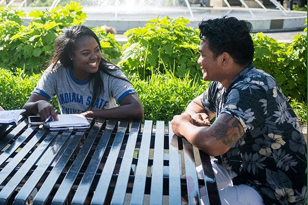 Two individuals sitting at a table outside next to a row of bushes. On the left is a Black female student wearing a grey-and-blue Indiana State University T-shirt. A notebook is in front of her and she holds a pencil. On the right is a male student with tanned skin. He wears a blue-and-white Hawaiian print shirt.