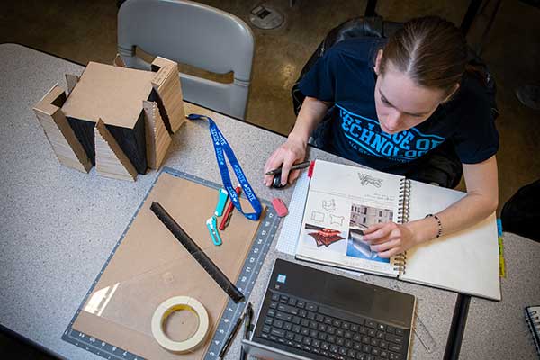 A student sits at a table, wearing a blue Bailey College of Technology T-shirt. A notebook is on the table and the student looks at a laptop. Cardboard, a ruler, and tape are to the left.  