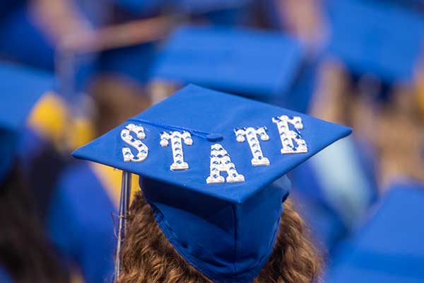 A graduating student wearing a cap and gown at her commencement ceremony at Indiana State University. The back of her blue graduation cap shows the word “STATE” in white and blue letters. 