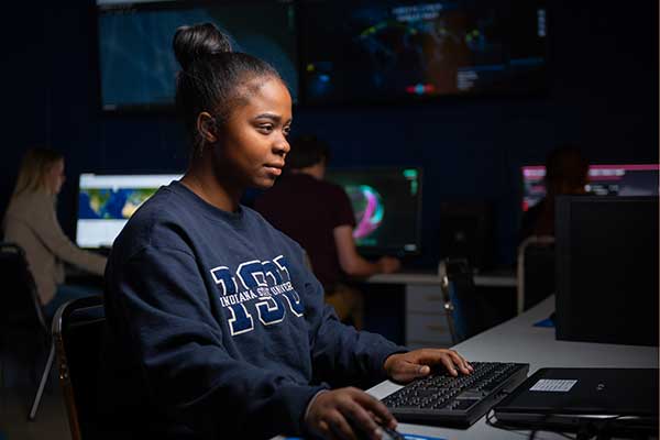 A Black graduate student wearing an Indiana State University sweatshirt works at her computer in a darkened Cyber/Intel Lab. 