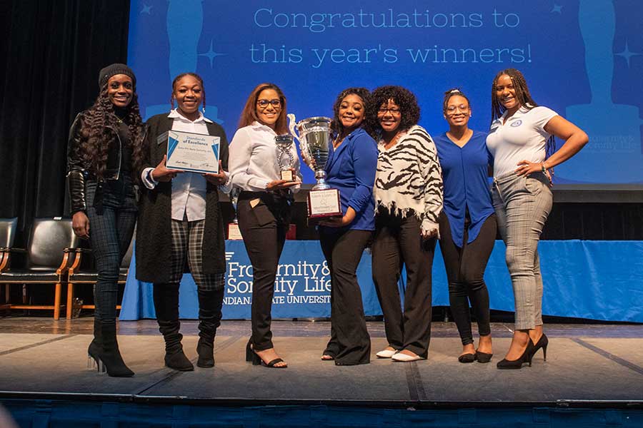 A group of seven Black female students celebrates on a stage after receiving an award for excellence. Two women in the middle hold up a large silver trophy, and the woman to their left holds up a framed certificate. 