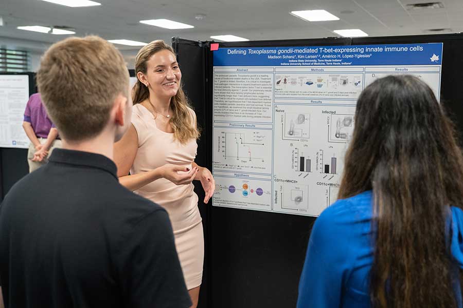 A smiling graduate student in a beige, short-sleeved dress with dark blonde hair explains her microbiology research poster to two other students. The poster features text, charts, graphs, and diagrams.  