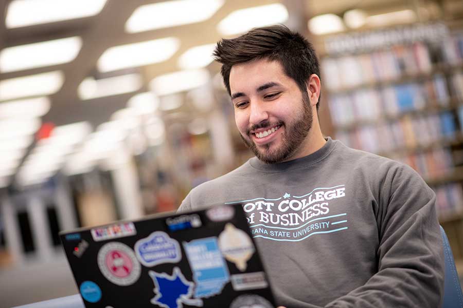 A smiling male student with black hair and beard sits in the Cunningham Memorial Library with an open laptop. He is wearing a grey, long-sleeved sweatshirt that says ‘Scott College of Business’ and ‘Indiana State University’ on it. The laptop has assorted stickers on the cover, including a blue Sycamore leaf. Books on shelves are visible, but out of focus, in the background.     