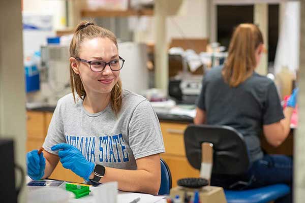 A White female student, with dark blonde hair in a ponytail, smiles in the direction of an unseen classmate as she works with test tubes in a laboratory classroom. She is wearing glasses, latex gloves, and a grey Indiana State sweatshirt with blue letters. 