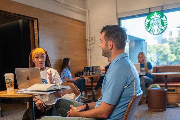 A Black female student in a white t-shirt with a blue decal, and wearing a yellow head scarf, sits behind a table at a Starbucks café with a Macbook open in front of her. To her left sits a White male with dark brown hair, wearing a light blue shirt. A paper notepad and a large open book sit in front of him. The Starbucks is logo is visible hanging above a large window behind them.   