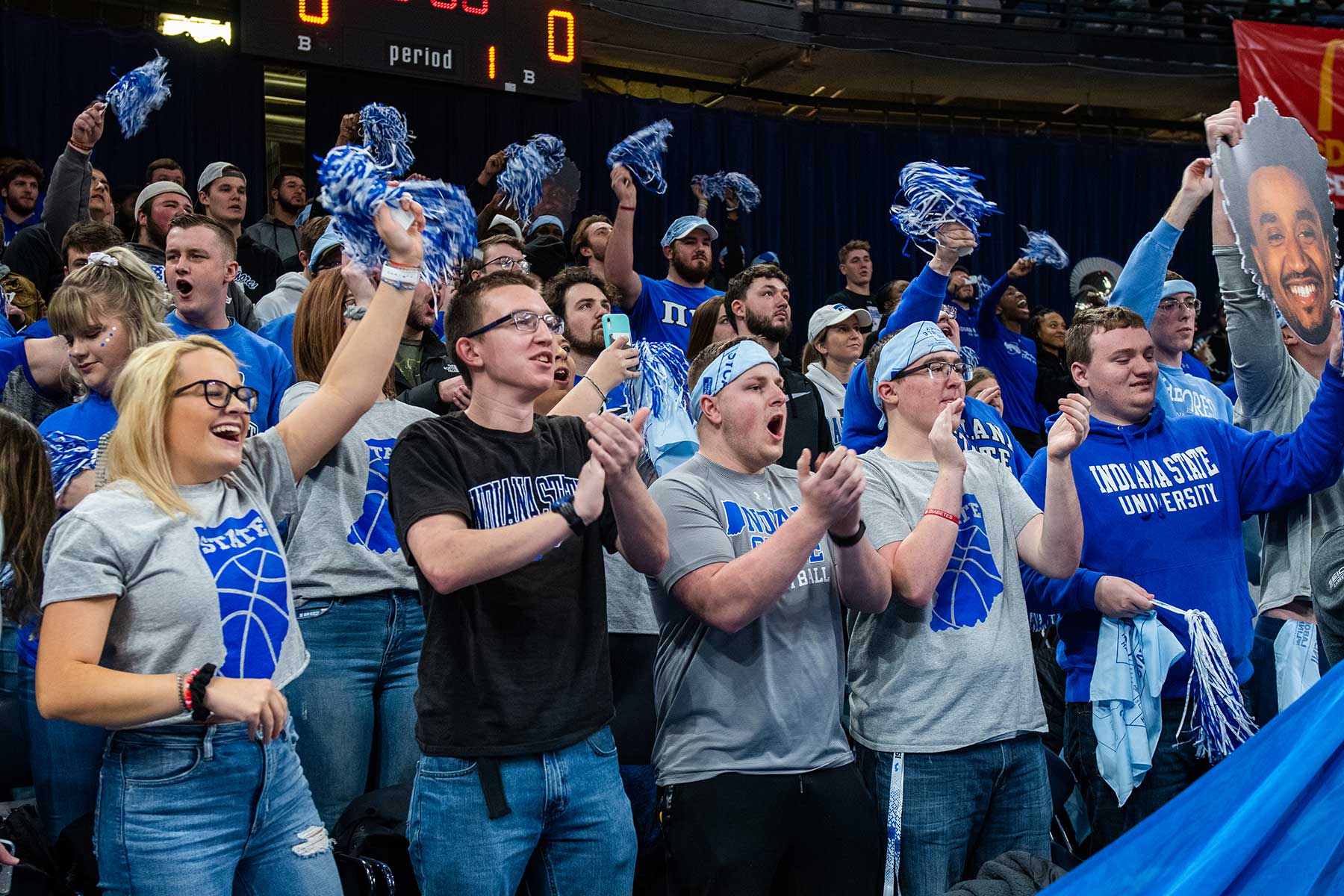 A large group of Sycamore fans, men and women, cheer for their team during a basketball game at the Hulman Center. They are clapping, cheering, holding blue and white mini-pompoms, and many are wearing light blue headbands. 
