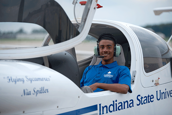 A Black male student sits in a small white airplane with Indiana State University printed on the side. He wears a blue Indiana State polo shirt and a headset.