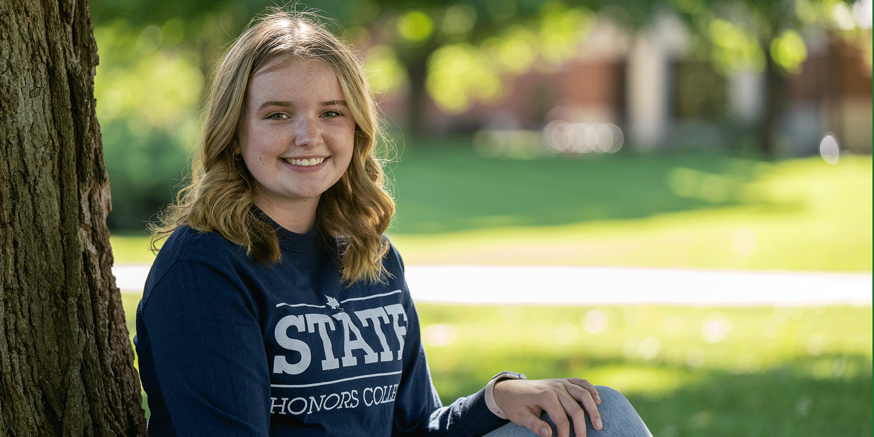 A female student with curly blonde hair sits with her back to a tree. She wears a blue long-sleeved STATE Honors College shirt and blue jeans. 