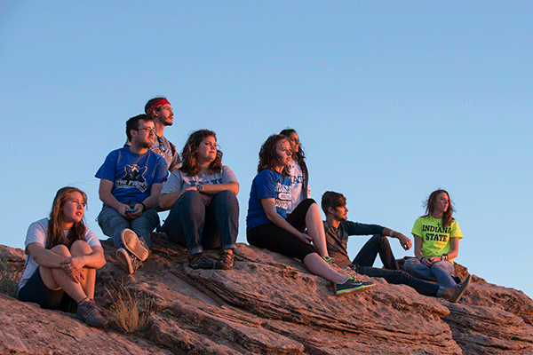 Eight Honors students, of mixed races, sit on a canyon rock, looking to the right. They wear a variety of blue, white, grey, and green clothing.